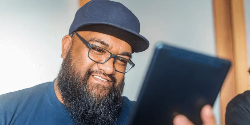 A bearded man wearing glasses and a navy cap smiles while looking at a tablet screen.