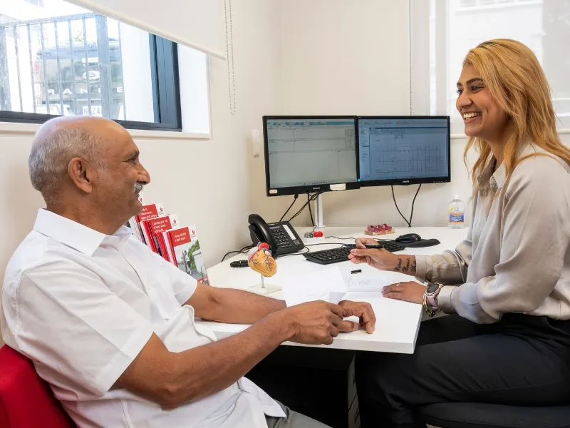 A doctor consults with an elderly male patient in a modern medical office, discussing heart health as visible on dual computer monitors displaying medical charts.