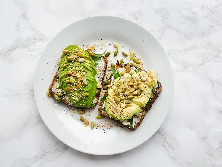 Two pieces of wholegrain toast topped with avocado and nuts and seeds on a white place and a white and gray marble surface.