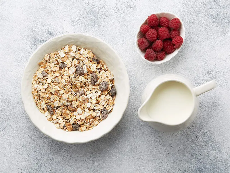 A top-down view of a bowl of breakfast oats in a white bowl on a marble tabletop. Alongside is a white jug of milk and a small with bowl of fresh raspeberries.
