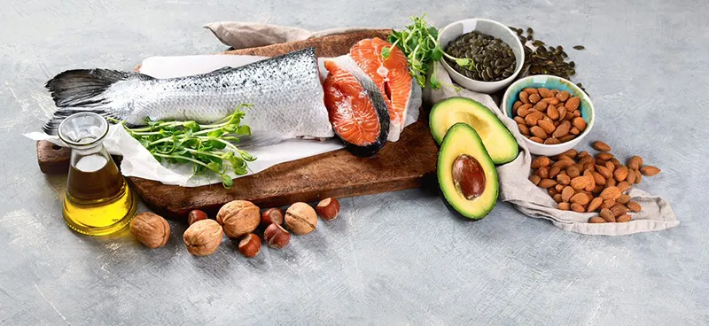 A display of foods on a wooden board. Olive oil in a glass jar, alongside salmon, various nuts and seeds plus avocado.