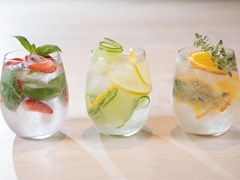 Three glass tumblers of infused water arranged in a row on an off-white surface with additional ingredients in the background.