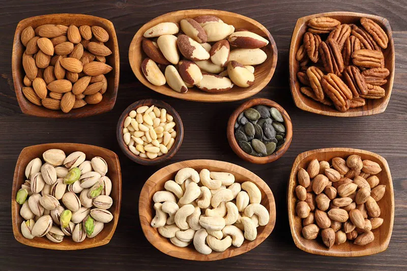 A top-down view of eight different types of nuts displayed in wooden bowls of differing sizes. They sit on a wooden table top.