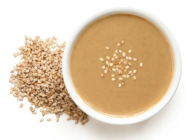 Tahini in a white ceramic bowl sprinkled with sesame seeds next to a pile of sesame seeds. 