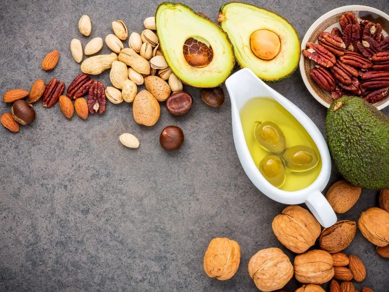 Assorted healthy fats including halved avocado, a jug of olive oil with olives, pecans, walnuts, almonds, pistachios, and various mixed nuts arranged on a dark countertop.