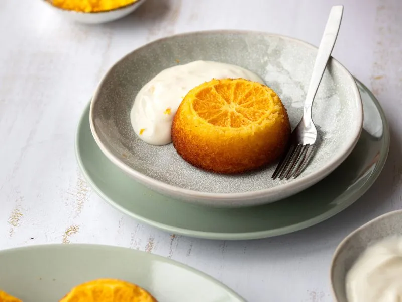 Individual orange upside-down cake dessert served on a small plate with creamy yogurt and a fork, placed on a light-colored surface.