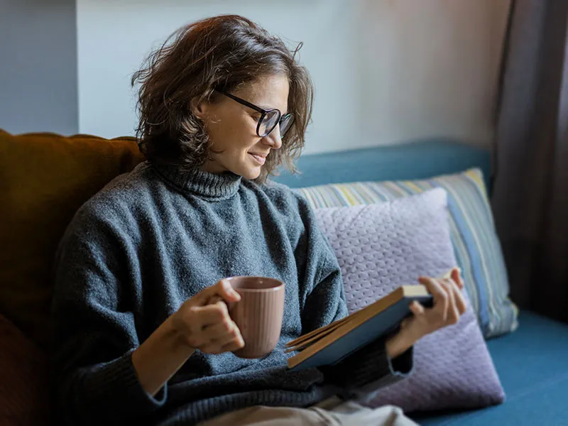 A smiling woman sits curled up on a sofa with a book and a hot drink.