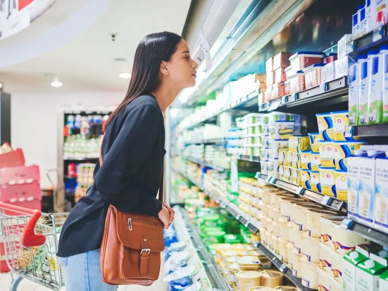 Woman standing in front of a refrigerated supermarket shelf, examining various packaged dairy and spread products with a shopping cart beside her.