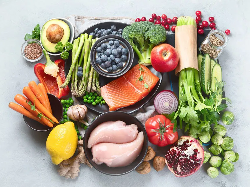 An assortment of low cholesterol foods displayed on a marble tabletop. Fruit, vegetables, nuts, seeds, salmon and chicken.