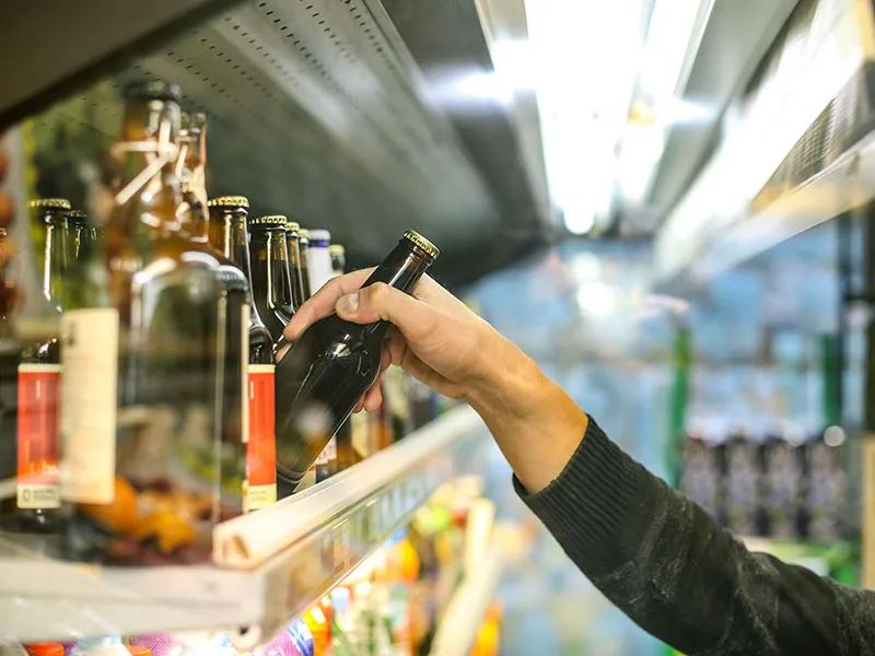 A shopper's hand is shown taking a bottle of beer from a shelf in a supermarket.