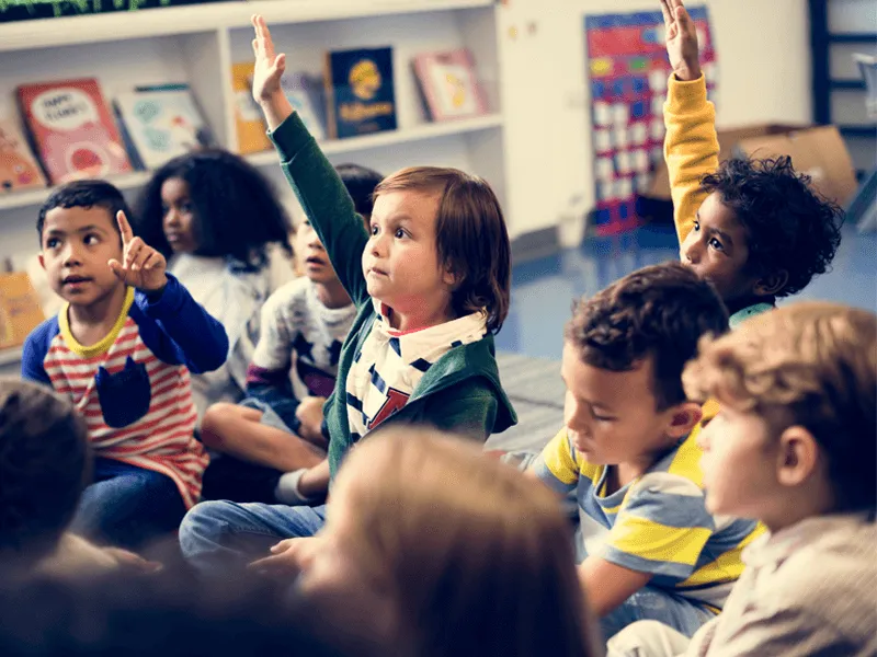 A group of young children sitting on the floor in a classroom during a group activity or lesson. Several children have their hands raised, eager to participate. They are dressed in casual clothing and are seated in front of a bookshelf filled with colorfu