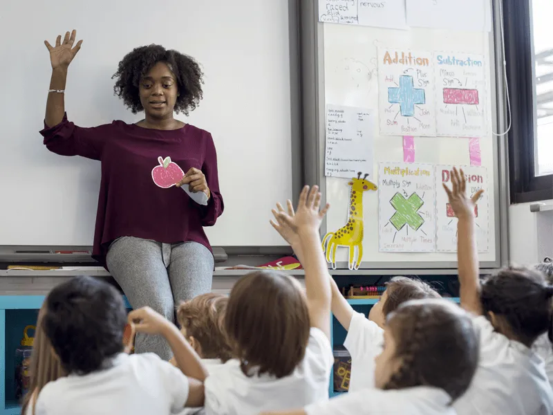 Teacher sitting at the front of a classroom holding a paper cut-out of an apple, with one hand raised, engaging with a group of young children. Several children, seated on the floor and facing the teacher, are raising their hands enthusiastically. Behind