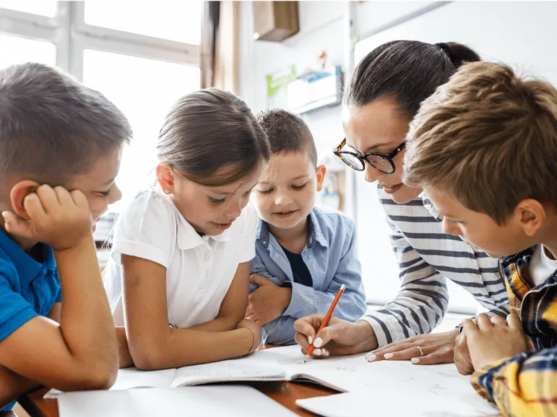 Teacher working with a group of four young children gathered around a table, helping them with a writing activity in an open, bright classroom.
