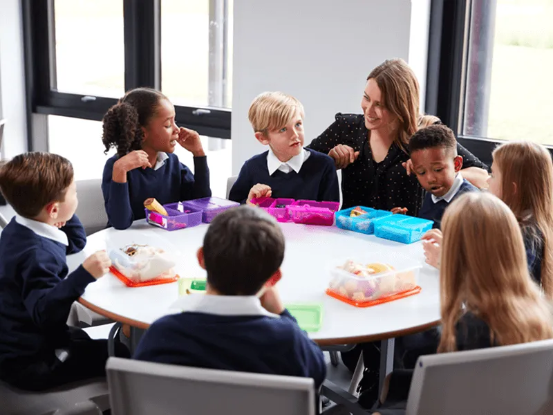 A group of young schoolchildren sitting around a circular table having lunch with a teacher. The children are wearing navy blue school uniforms with white collars. They are engaged in conversation and eating from colorful lunchboxes. The teacher is smilin