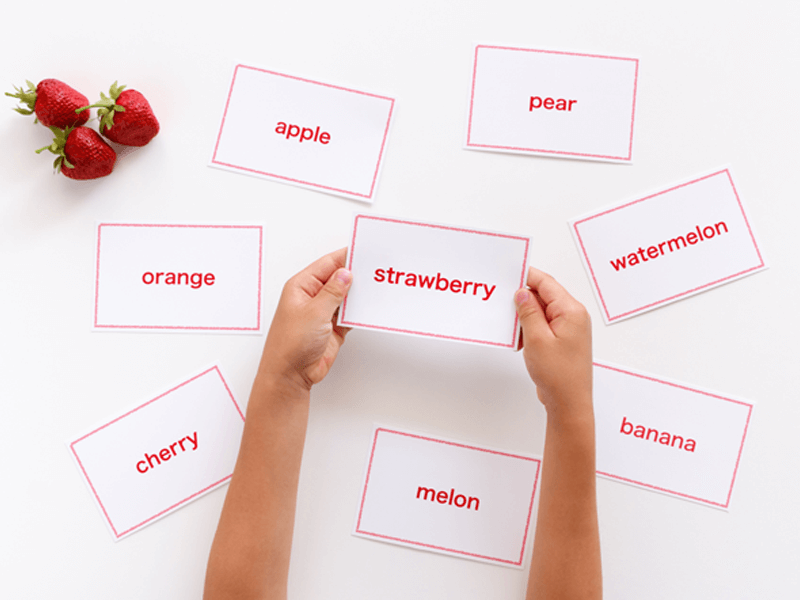 Child's hands holding a flashcard labeled 'strawberry', surrounded by other flashcards with names of fruits including apple, pear, orange, watermelon, cherry, melon, and banana, on a white background with fresh strawberries placed to the side.