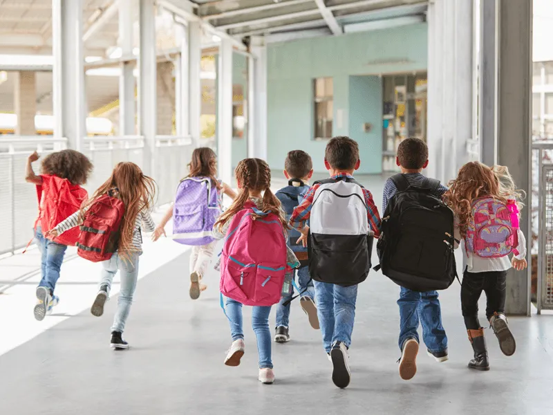 Group of young schoolchildren running down a covered outdoor walkway, seen from behind. The children are wearing casual clothes and carrying colorful backpacks. The scene suggests the end of the school day or excitement for recess or a break. The walkway